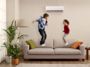 Two children jumping on a beige couch in front of a beige wall where a mini split heat pump head is affixed above them.