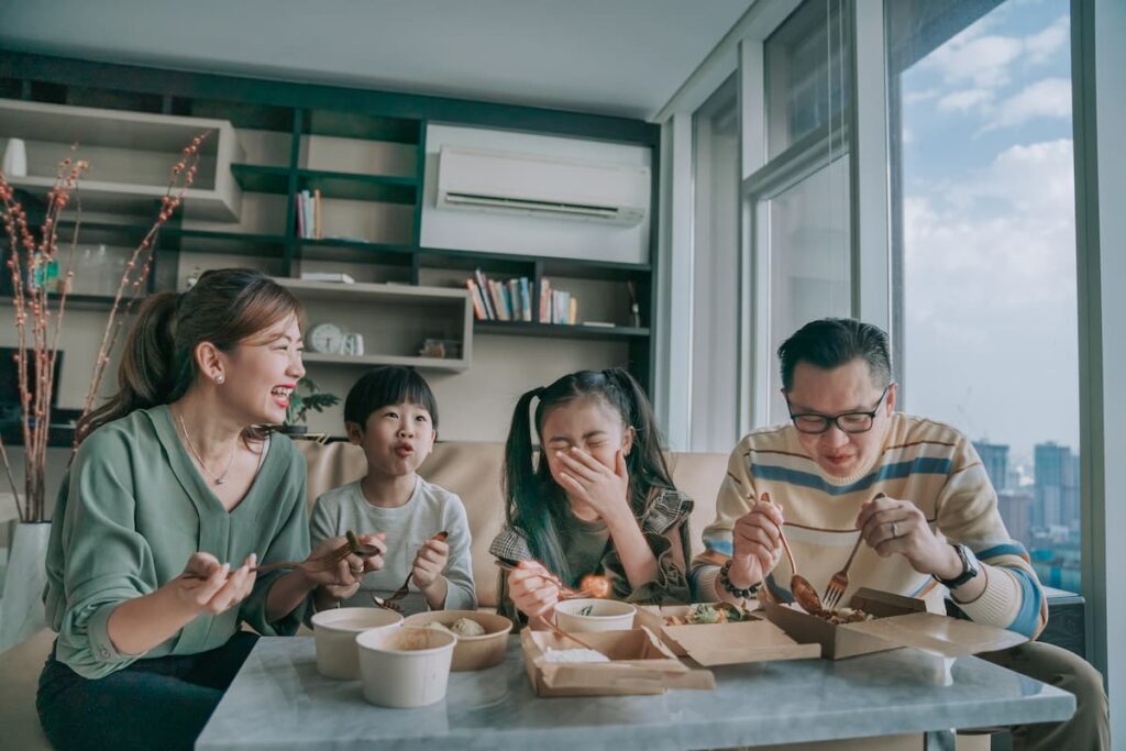 A young Asian family is sitting side by side on a couch eating a meal and laughing.