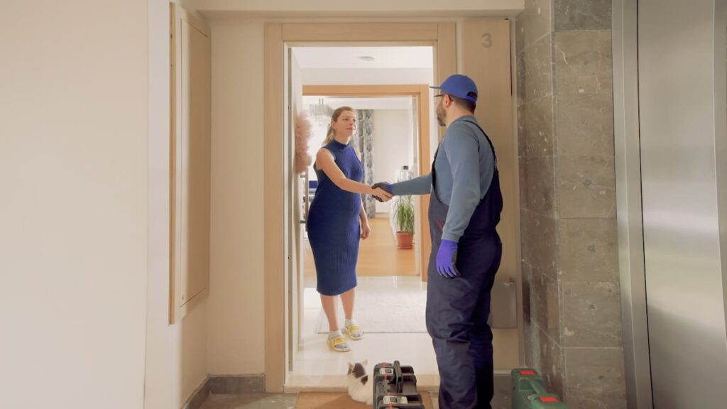 A white woman in a blue dress is standing in a doorway, shaking hands with a man wearing gloves and overalls with a toolbox at his feet.