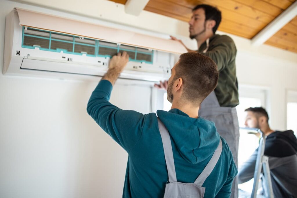 Two bearded men in overalls are doing work on an open ductless heating and cooling unit on the wall.