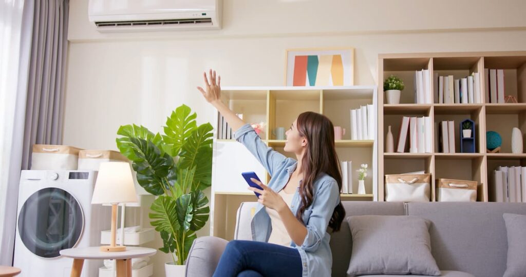 A woman with long brown hair is sitting on a couch; she is raising her hand toward the cooling unit behind her to feel the cool air.