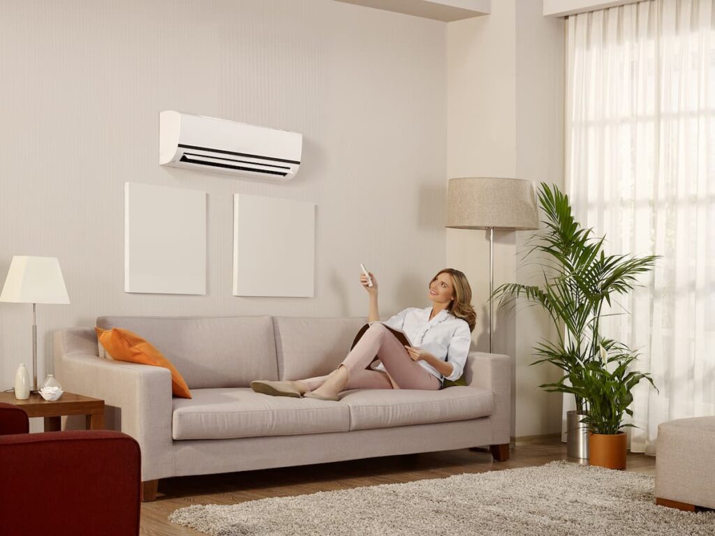 A white woman with blonde hair is sitting on a light brown couch. She is looking up at a minisplit unit and pointing a remote at it while smiling